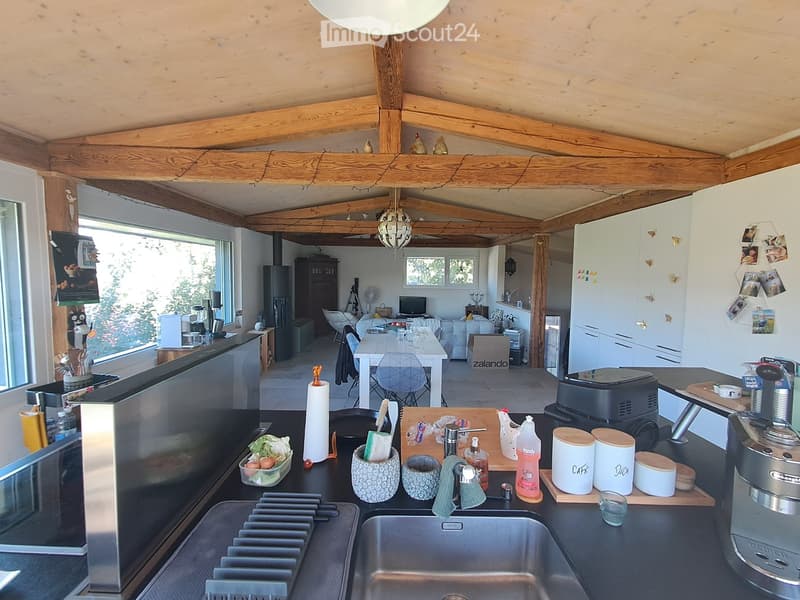 Open-plan kitchen and living area with exposed wooden beams, modern appliances, and a dining table with chairs. The kitchen features a sink, coffee maker, and various utensils, while the living area includes a sofa, laptop, and a fan.