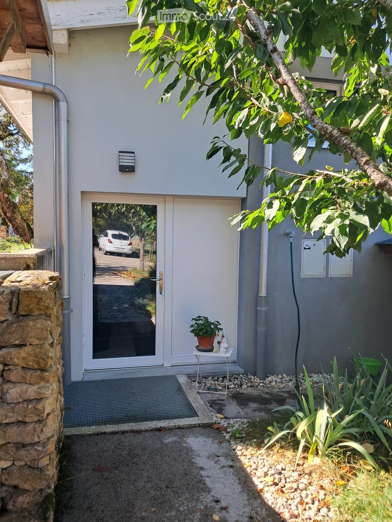 Entrance area of a house with a small table holding a potted plant and decorative items, surrounded by greenery and a stone wall.