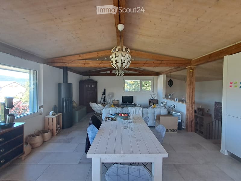 Open-plan living and dining area with exposed wooden beams, a large wooden dining table, white sofa, TV, and various decorative items.