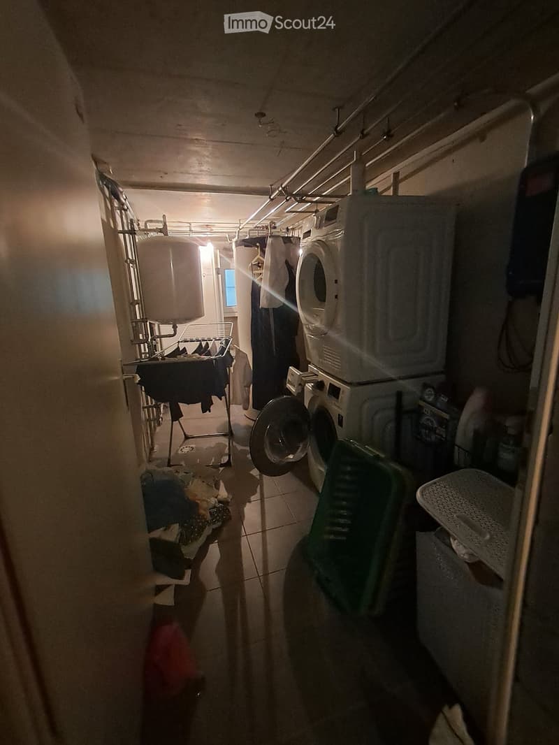 A narrow hallway with a washing machine and dryer stacked, clothes hanging on a rack, and various items scattered on the floor.