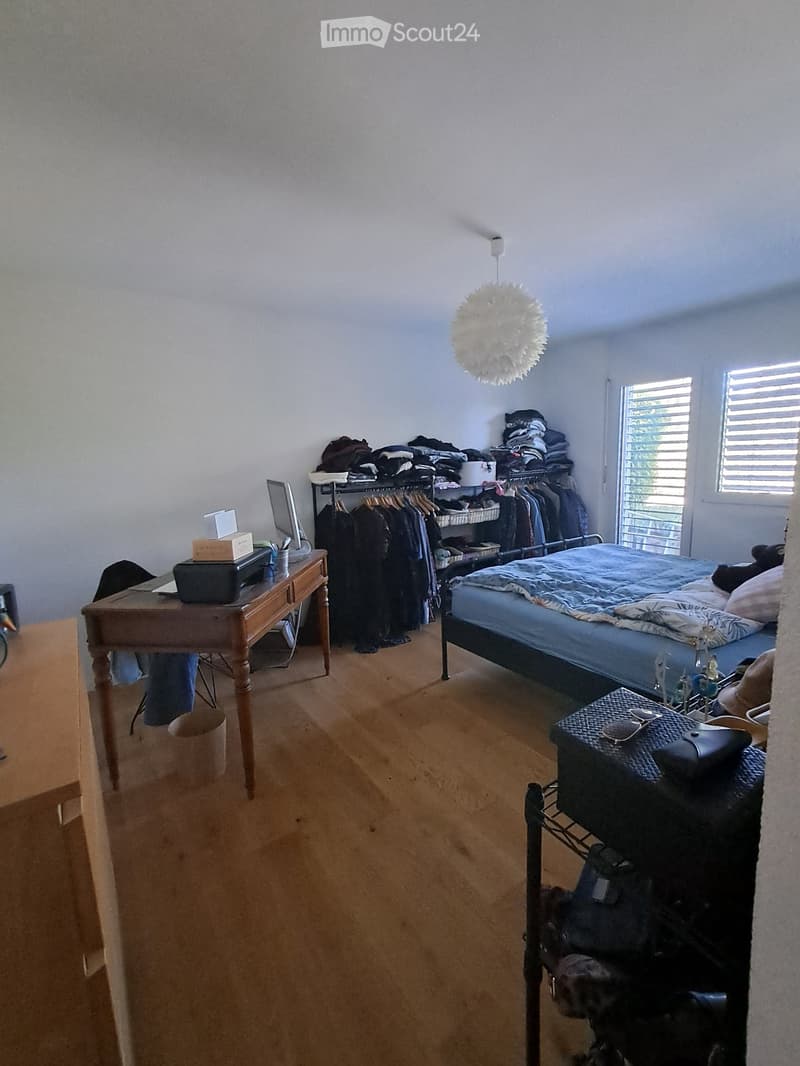 Bedroom with wooden floor, bed, desk, wardrobe, and clothing items. Features a hanging lamp and a window with blinds.