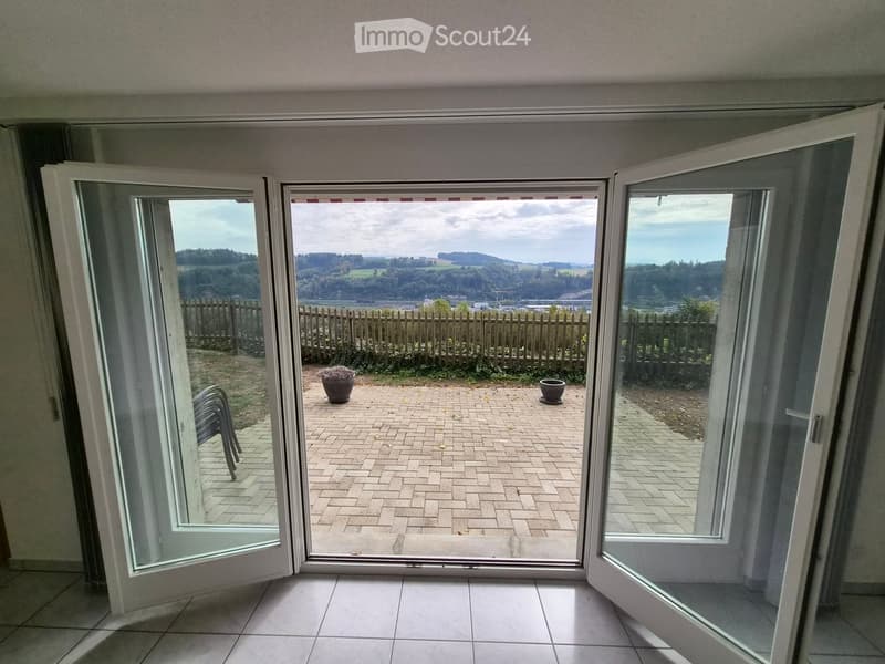 Open sliding doors lead to a brick-paved terrace with a scenic view of a valley and hills, featuring a wooden fence and potted plants.