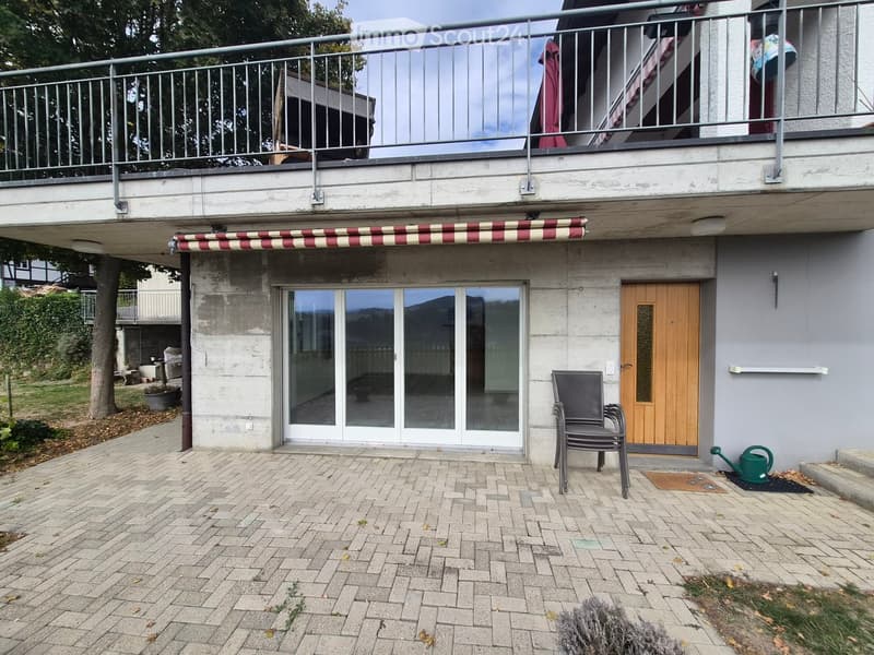 Brick-paved outdoor area with a black metal chair, green watering can, and a wooden door leading to the house. A large sliding glass door is visible, and a striped awning extends from the upper level.