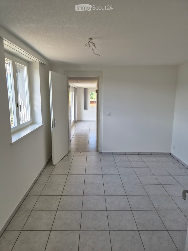 Empty hallway with tiled flooring, white walls, and a window on the left side. The hallway leads to another room with visible doors and a light fixture on the ceiling.