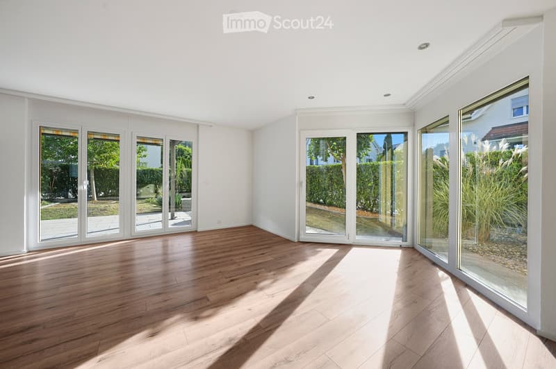 Bright, empty room with wooden flooring and large glass sliding doors leading to a garden. The space is clean and minimalistic with white walls and recessed lighting.