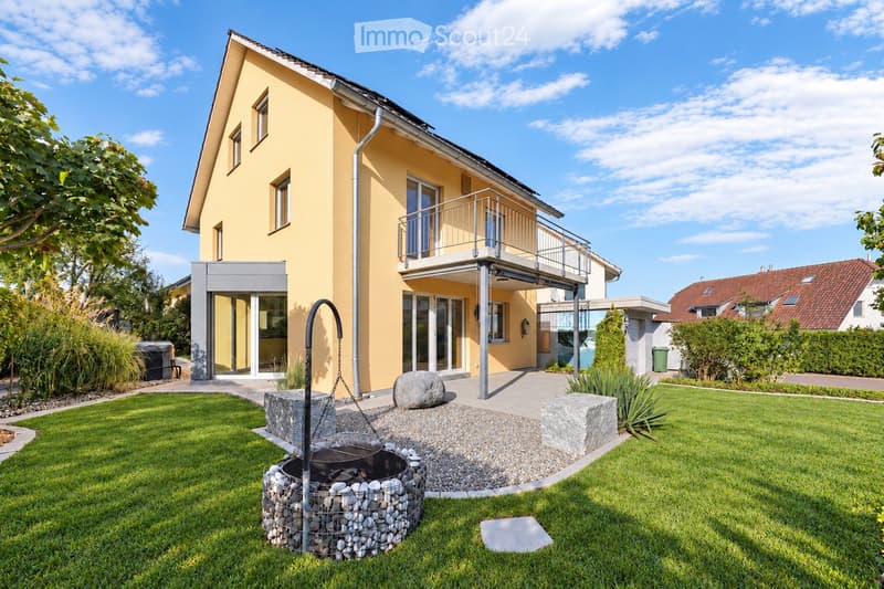 Two-story yellow house with a modern design, featuring a balcony, large windows, and a well-maintained garden with a stone fire pit.