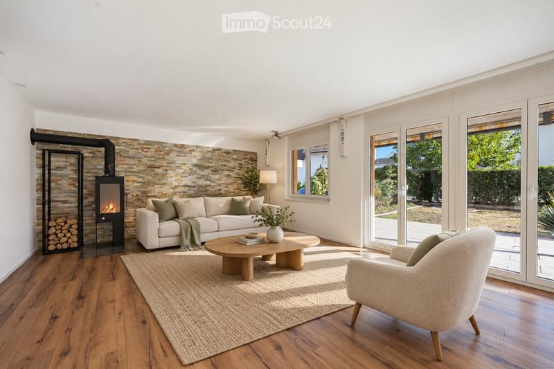 Modern living room with a white sofa, wooden coffee table, armchair, fireplace, and large windows overlooking a garden.