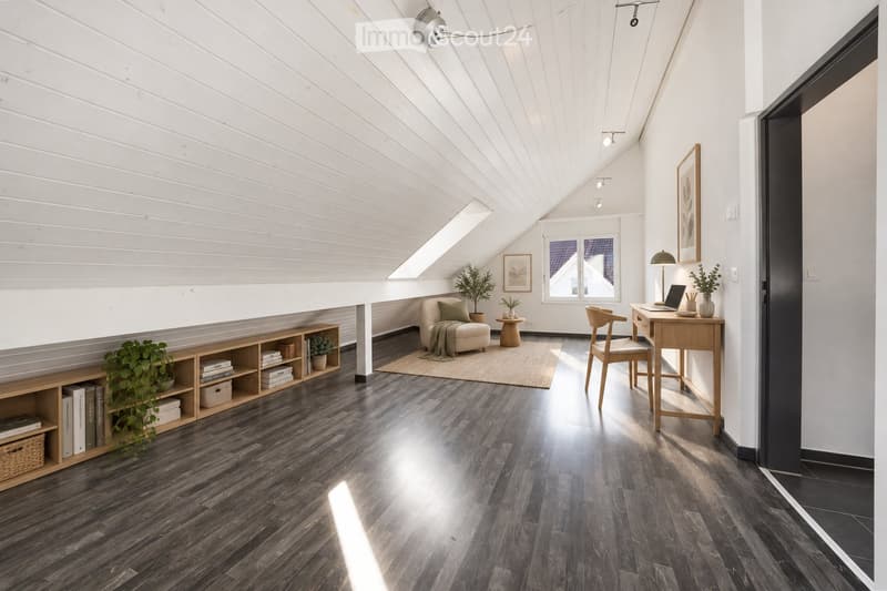 Bright, modern attic room with wooden floors, slanted white ceiling, large windows, and skylight. Features a sofa, desk with laptop, chair, shelf with books and plants, and a small round table with a plant.