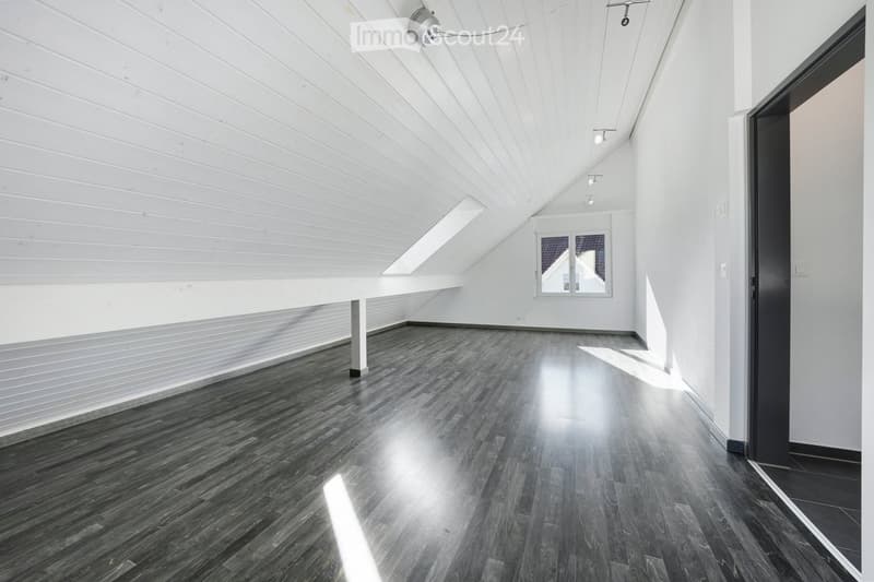 Empty room with dark wooden flooring, white walls, and a slanted white ceiling. Features a large window and a black door frame.