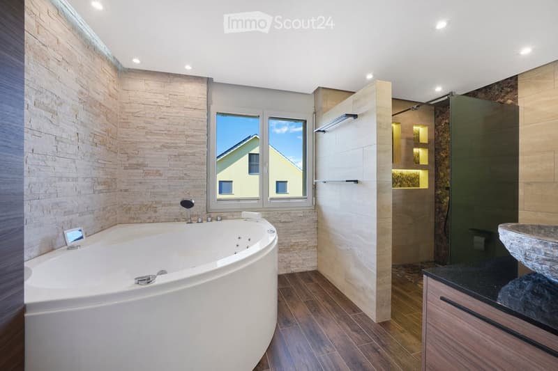 Modern bathroom with a large, curved bathtub, a stone sink, a glass-enclosed shower stall, and wooden flooring.