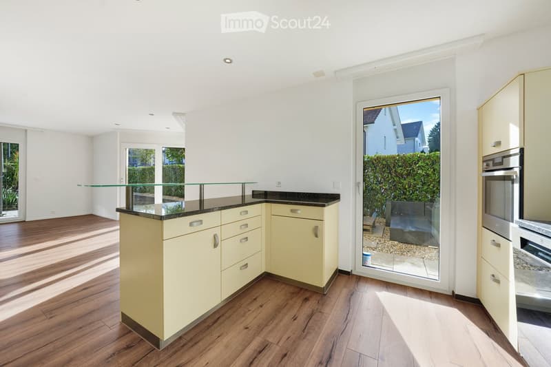 Modern kitchen with light yellow cabinets, dark countertops, and a glass island. Features include wooden flooring, large windows, and built-in appliances.