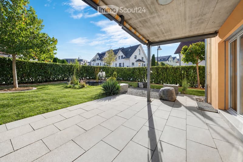 A modern patio area with large concrete tiles, a well-maintained lawn, a small fountain, and surrounding greenery, including trees and shrubs. The patio is covered by a sleek, contemporary roof structure.