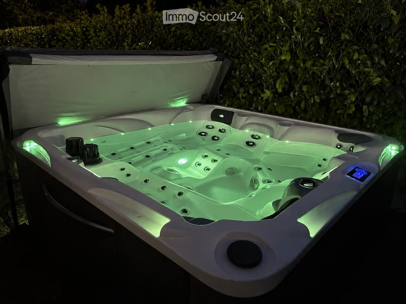 A modern hot tub with green LED lighting, surrounded by lush green foliage, featuring multiple jets and a control panel.