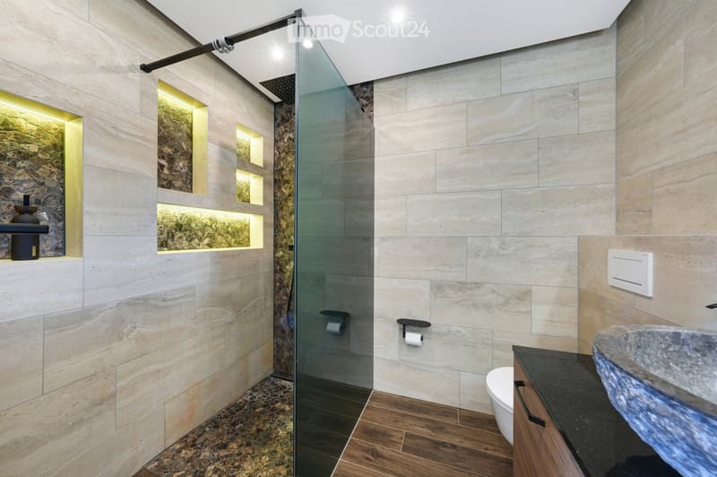 Modern bathroom with marble tiles, glass shower enclosure, wooden floor, toilet, sink, and decorative lighting.