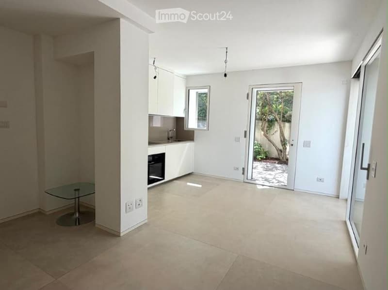 Open-plan unfurnished room with tiled flooring, white walls, built-in kitchen cabinets, and large sliding glass doors leading to a balcony area.