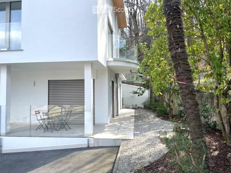 Modern two-story white building with a glass balcony and outdoor seating area, surrounded by trees and a gravel pathway.