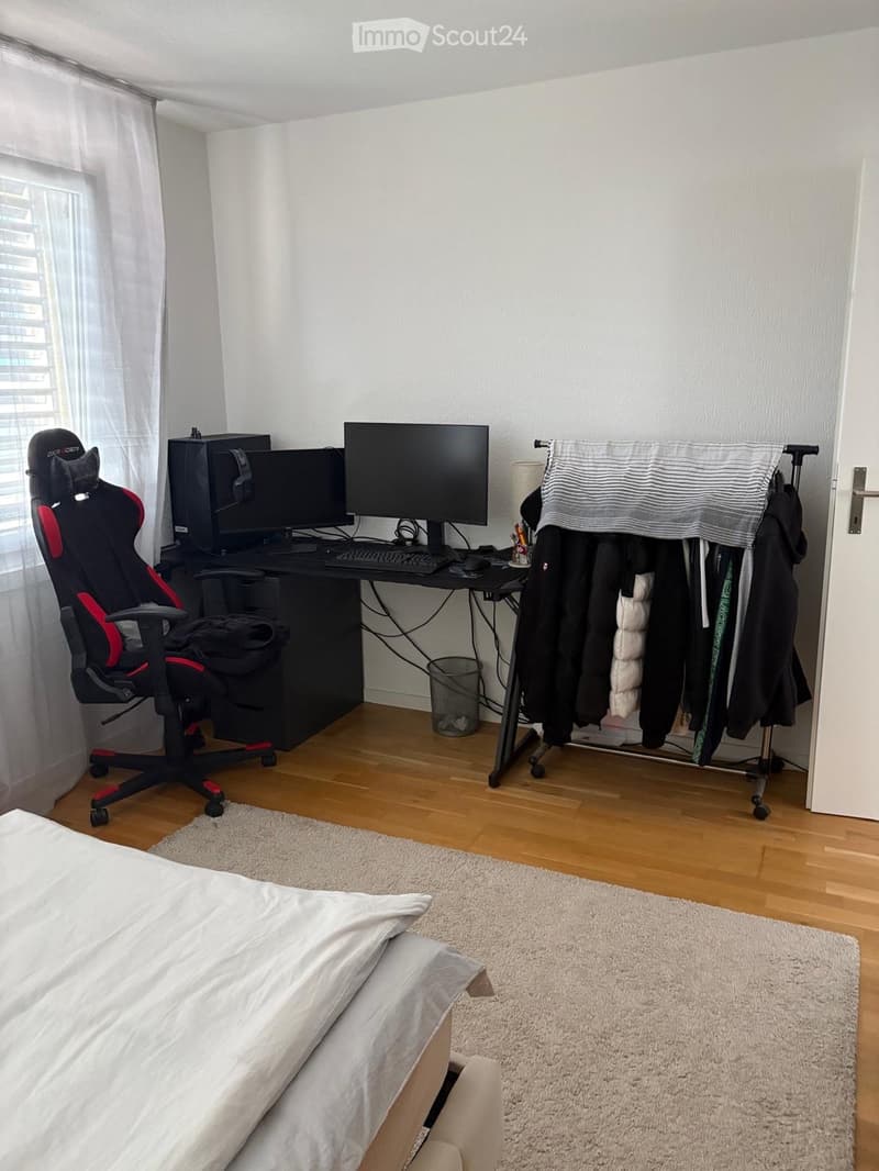 A furnished room with a bed, desk, gaming chair, computer setup, and a clothing rack. The room features wooden flooring and white walls.