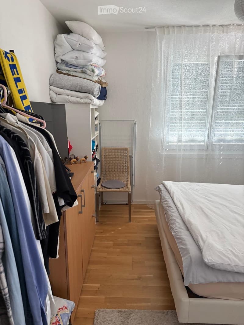 A small, neatly arranged bedroom with a bed, a wooden wardrobe, a shelf with folded blankets, a wicker chair, and a window with sheer curtains.