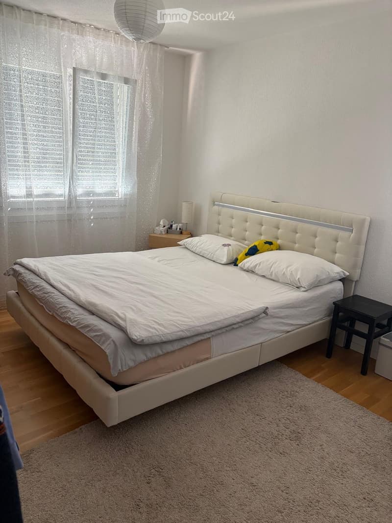 A modern bedroom with a large bed, white bedding, a tufted headboard, a nightstand with a lamp, and a small black stool.