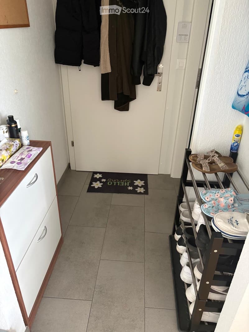 Hallway with tiled floor, white walls, coat rack with jackets, shoe rack with various shoes, small shelf with toiletries, and a doormat with text "HELLO".