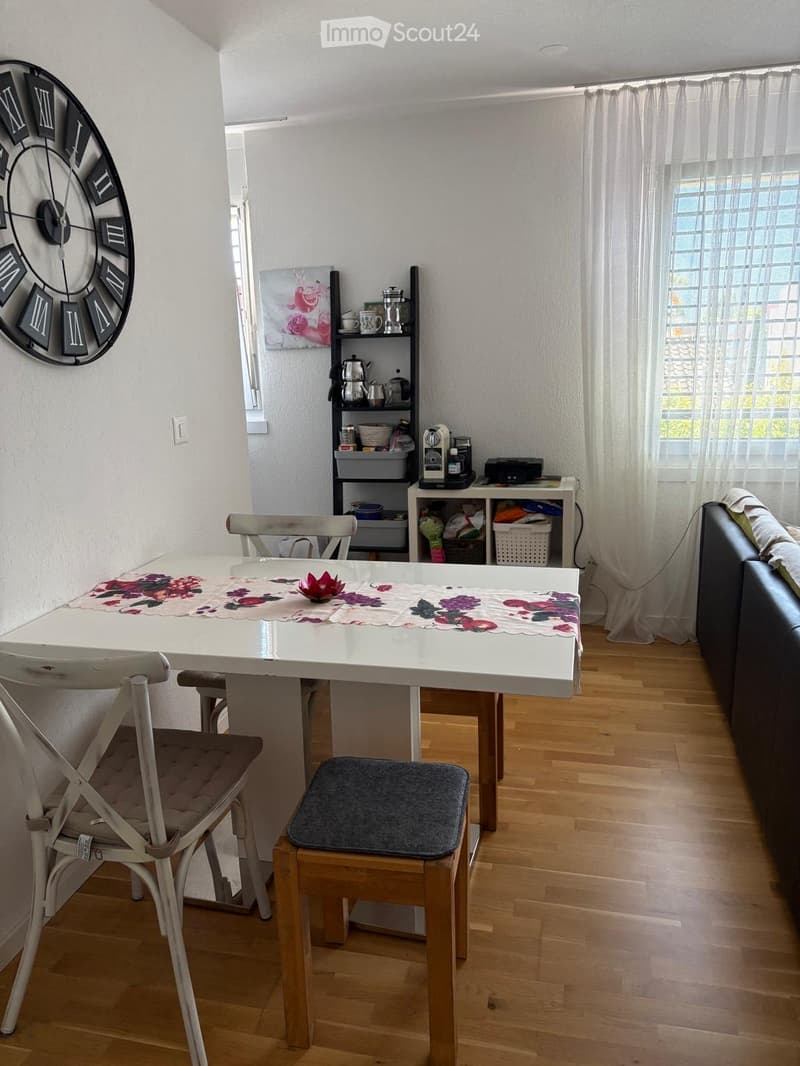 A cozy living and dining area with a white dining table, chairs, a sofa, shelves with kitchen items, and a large wall clock.