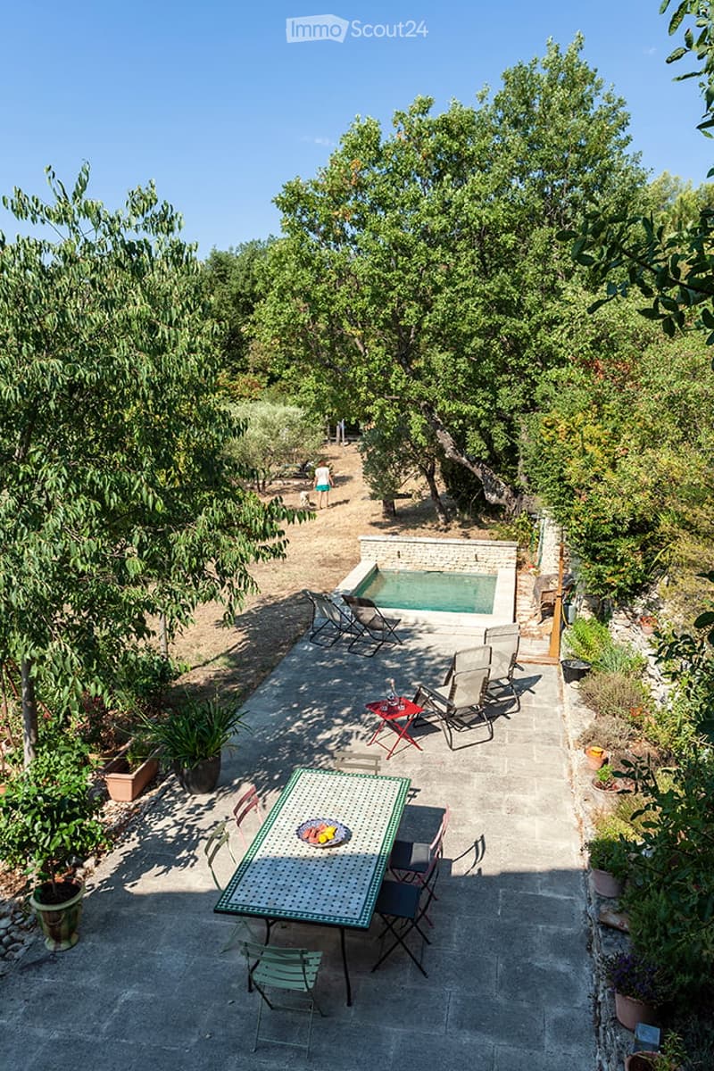 Outdoor patio with a dining table, chairs, and a swimming pool surrounded by trees and greenery.