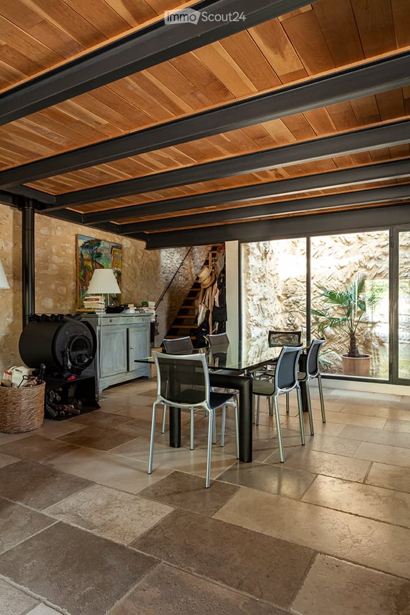 Open-plan dining area with exposed wooden ceiling beams, stone walls, and a large dining table surrounded by modern chairs. Features a fireplace, a cabinet with a lamp, and a potted plant near the large windows.