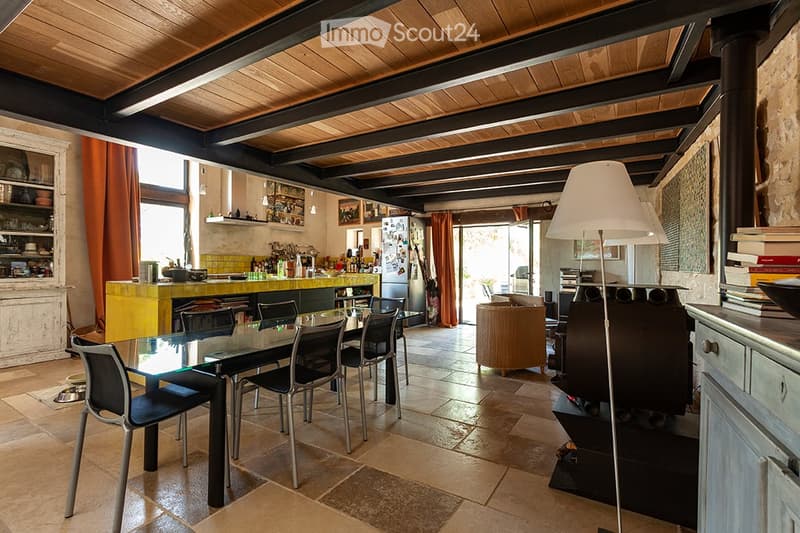 Open-plan living and dining area with exposed wooden beams, a glass dining table with chairs, a sofa, a floor lamp, and a kitchen area with a counter, refrigerator, and shelves.