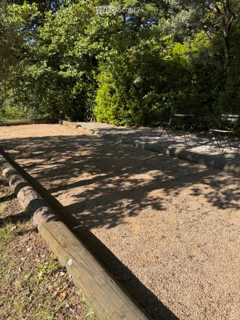 Outdoor area with gravel ground, wooden bench, small table and chairs, surrounded by trees and greenery.