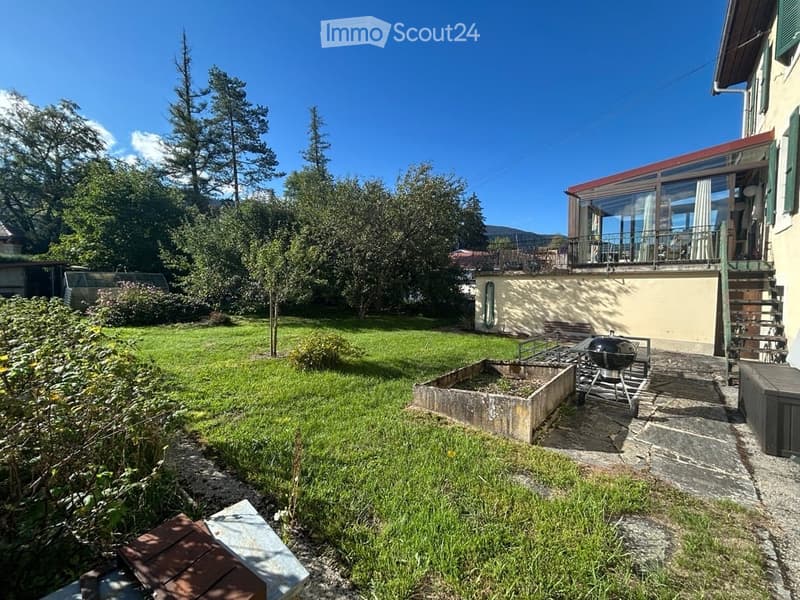 Well-maintained garden with a lawn, trees, a raised garden bed, and a barbecue. The house features a balcony with large windows and a staircase leading to the garden.