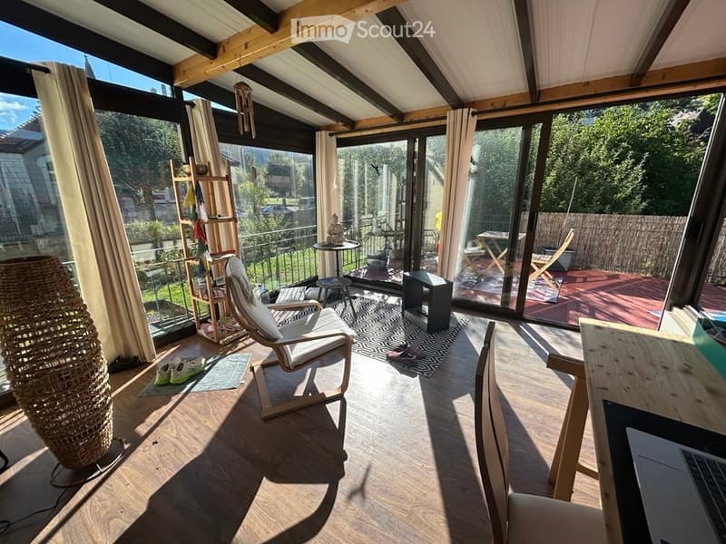 Bright, airy room with wooden flooring, large windows, and a balcony. Features a white chair, small table, stool, laptop, and decorative items.