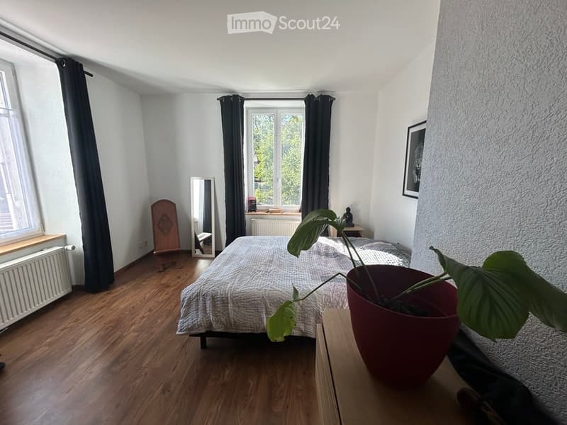 Cozy bedroom with wooden flooring, a neatly made bed, a large leafy plant in a pot, a mirror, and a small nightstand with a lamp.