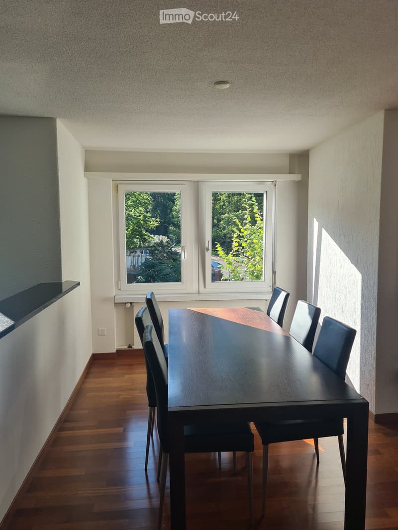 Dining room with a dark wooden dining table and matching chairs, large windows with a view of greenery, wooden flooring, and white walls.