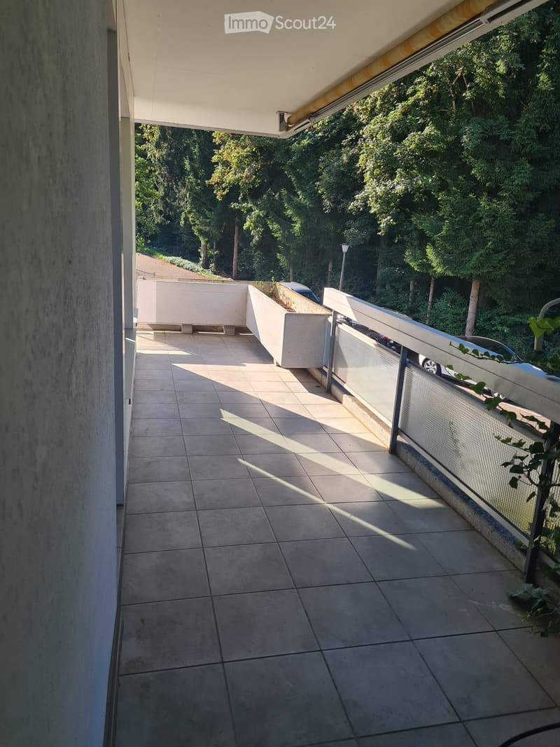 Balcony with tiled floor, modern railing, and a view of greenery and parked cars.