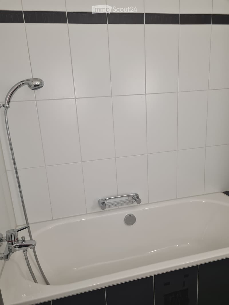 Modern bathroom with a white bathtub, chrome faucet, showerhead, and handrail. The walls are tiled in white with a black accent border.