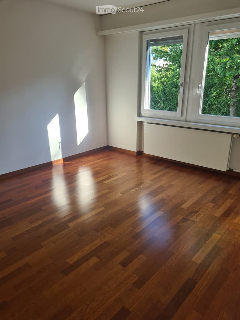 Empty room with wooden flooring, white walls, and large windows with white frames. A radiator is visible beneath the windows.