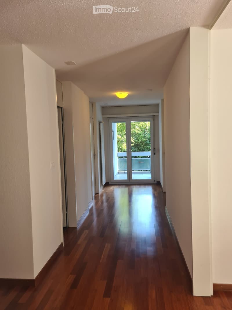 A long, narrow hallway with polished wooden flooring, white walls, and recessed lighting. The hallway leads to a glass door at the end, offering a view of a balcony area with outdoor furniture.