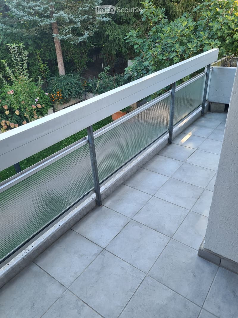 Balcony with modern glass railing, tiled floor, and lush greenery in the background.