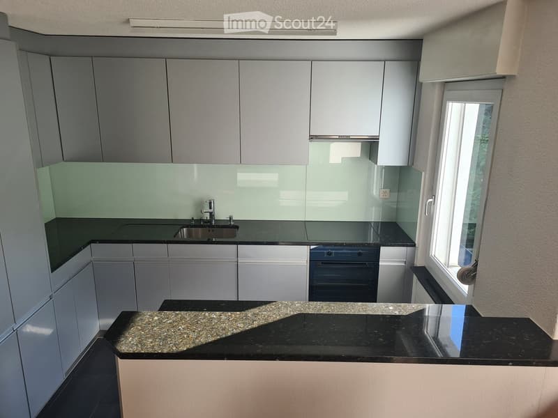 Modern kitchen with white cabinets, black countertops, a sink, and an oven. Features a large window with blinds and a granite island.