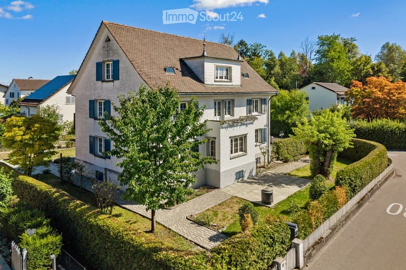 A two-story house with a brown tiled roof, white exterior, and multiple windows with shutters. The house is surrounded by a well-maintained garden with trees, shrubs, and a paved pathway leading to the entrance.