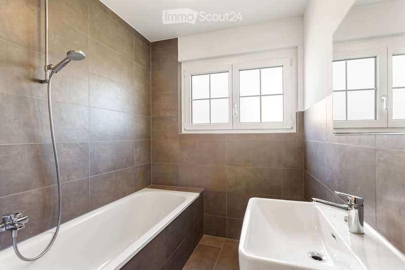 Modern bathroom with a bathtub, sink, and showerhead. The walls are tiled in brown and grey, and there are two windows providing natural light.