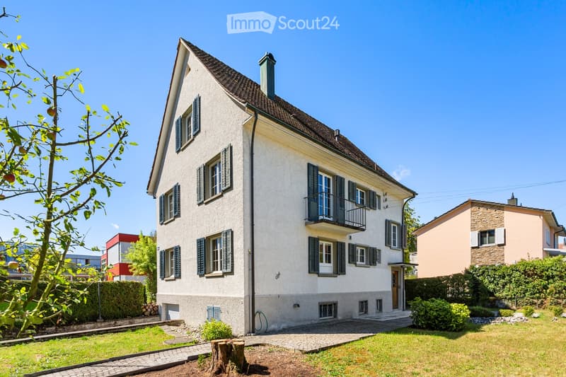 Two-story house with white exterior, dark shutters, and a balcony on the second floor. The house has a well-maintained garden with a tree, shrubs, and a paved driveway.