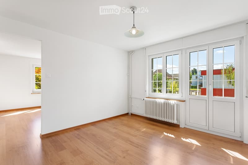 Empty room with wooden flooring, white walls, large windows, radiator, and a ceiling light fixture.