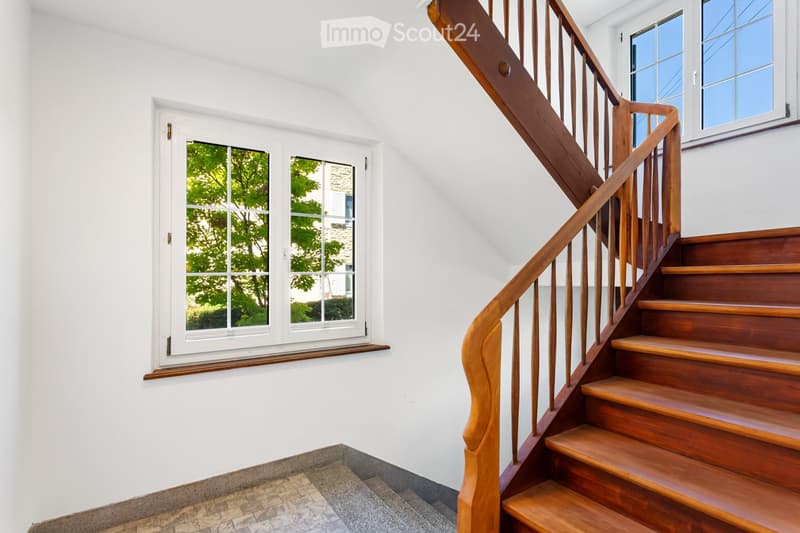 Wooden staircase with handrail and white walls, featuring a window with a view of greenery outside.
