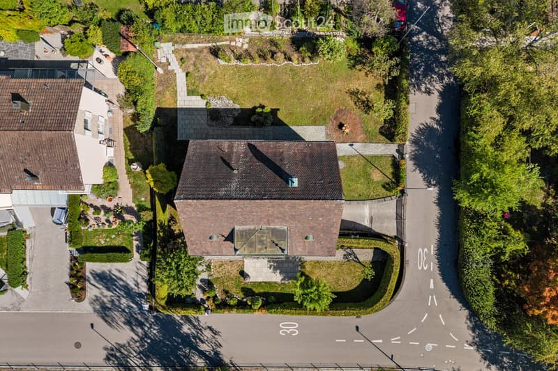 Aerial view of a two-story house with a tiled roof, surrounded by a well-maintained garden and driveway. The house features multiple windows and a small balcony area on the upper level.