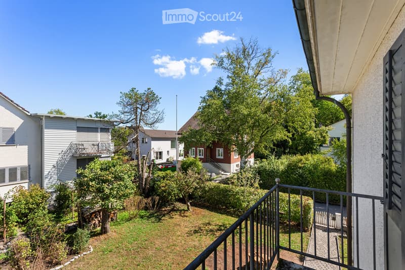 Balcony overlooking a garden area with trees, shrubs, and neighboring houses in the background.