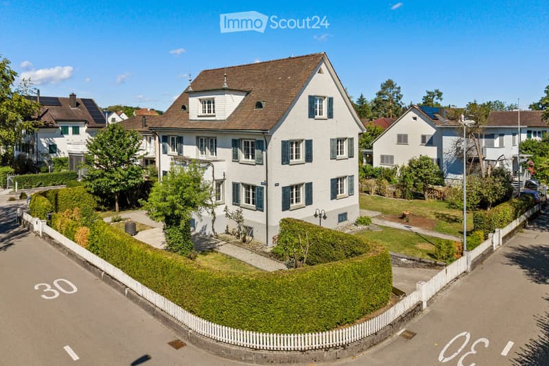 A two-story white house with blue shutters, surrounded by a well-maintained garden and white picket fence. The house features multiple windows and a pitched roof with dormer windows.