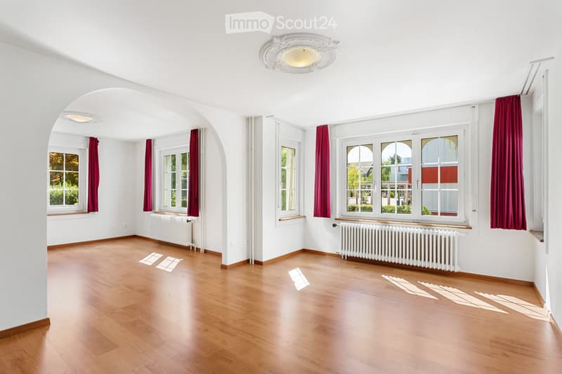 Empty room with wooden flooring, white walls, large windows with red curtains, radiators, and a decorative ceiling light fixture.