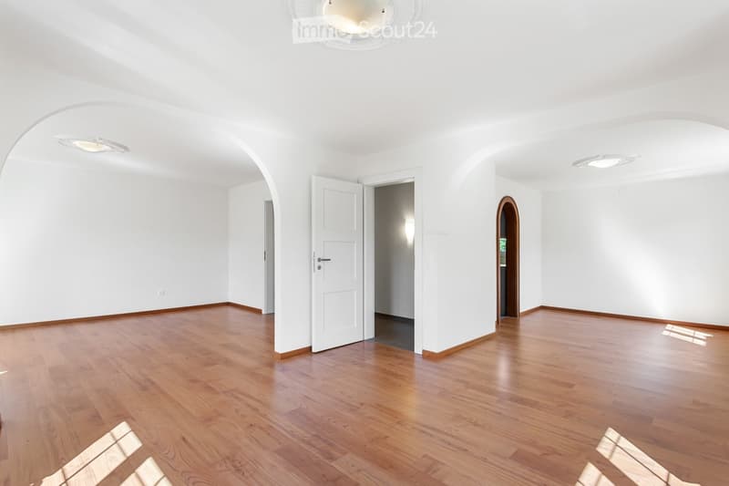 Empty room with wooden flooring, white walls, and several doors leading to other spaces. The room is well-lit with recessed ceiling lights.