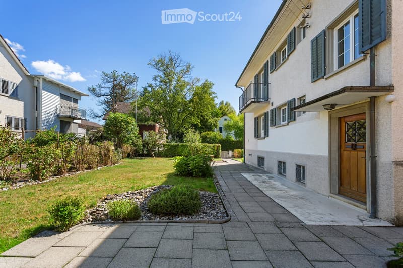 A well-maintained garden area with a paved pathway leading to a house entrance, surrounded by greenery and shrubs.